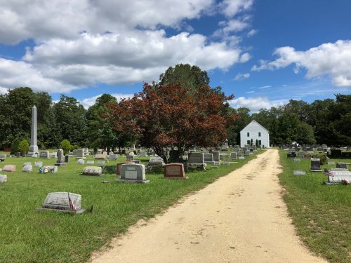 A peaceful cemetery with gravestones, a vibrant tree, and a white building under a blue sky with scattered clouds.