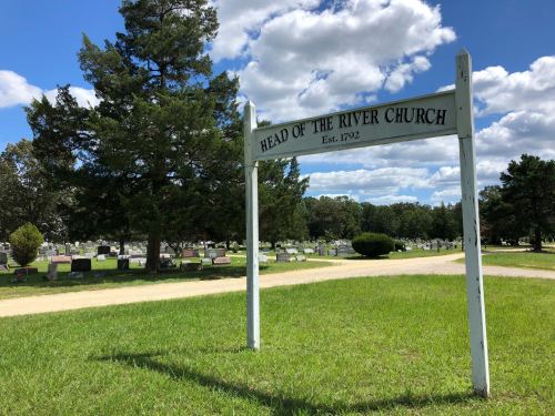 Sign for Head of the River Church, established in 1792, with a cemetery and trees in the background under a blue sky.
