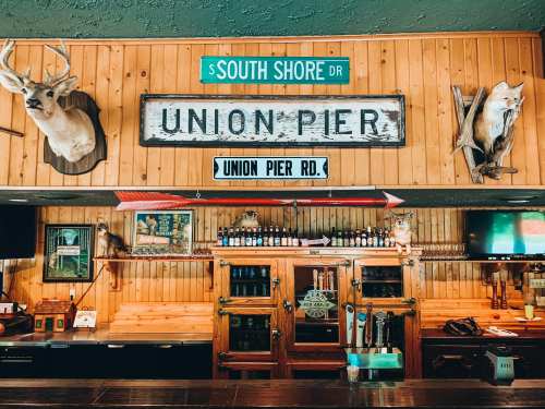 A rustic bar with wooden walls, vintage signs, and mounted animal heads decor. Bottles line the shelves behind the counter.