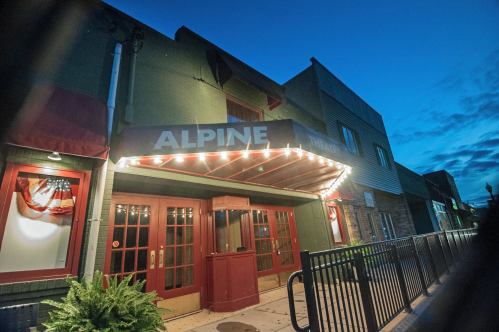 The Alpine building at dusk, featuring a marquee and bright lights, with a welcoming entrance and decorative plants.
