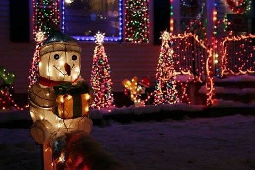A festive scene featuring a lit snowman holding a gift, surrounded by colorful Christmas lights and decorated trees.