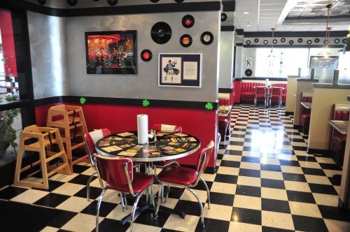 A retro diner interior with checkered floors, red booths, and a round table decorated with vinyl records on the walls.