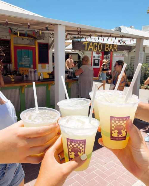 A group of people toasting with colorful drinks in front of a taco bar on a sunny day.