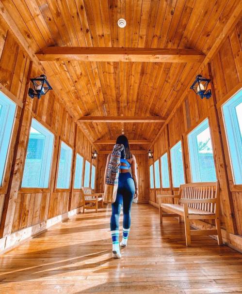 A person walks down a wooden hallway with large windows, wearing athletic clothing and carrying a bag over their shoulder.