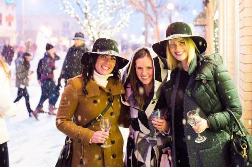 Three women in winter attire smile together in a snowy outdoor setting, with festive lights and a crowd in the background.