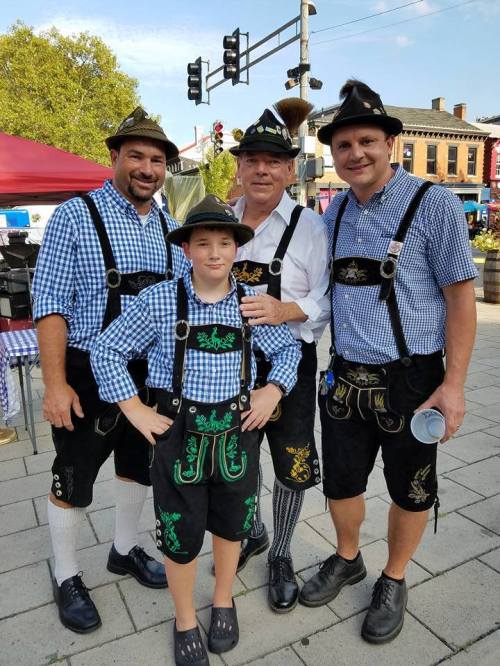 Four people in traditional Bavarian attire, including lederhosen and hats, pose together at an outdoor event.