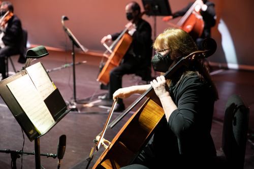 A masked cellist plays on stage, with other musicians in the background, all seated and focused on their music.