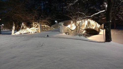 A snow-covered landscape at night featuring a bridge adorned with festive lights and trees in the background.