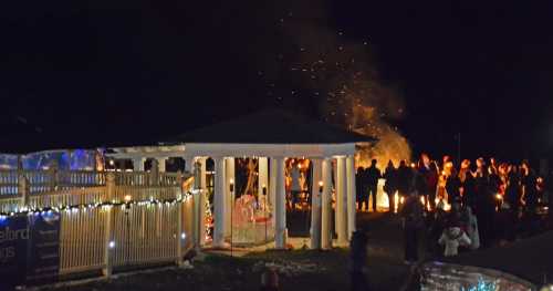 A nighttime gathering with people around a fire, illuminated by festive lights and a warm atmosphere.