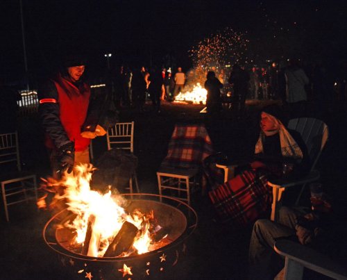 A group of people gather around a large fire pit at night, enjoying warmth and conversation in a festive atmosphere.