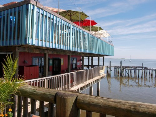 A colorful waterfront restaurant with a blue upper deck and red lower level, overlooking calm waters and a sailboat.