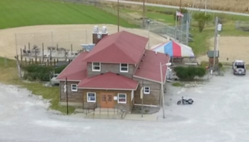 Aerial view of a small building with a red roof, surrounded by a baseball field and grassy areas.