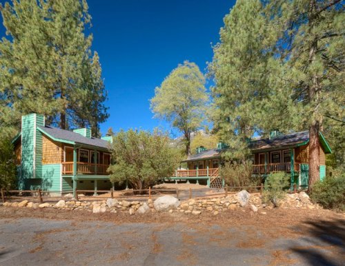 Two rustic cabins surrounded by tall trees and rocky landscaping under a clear blue sky.