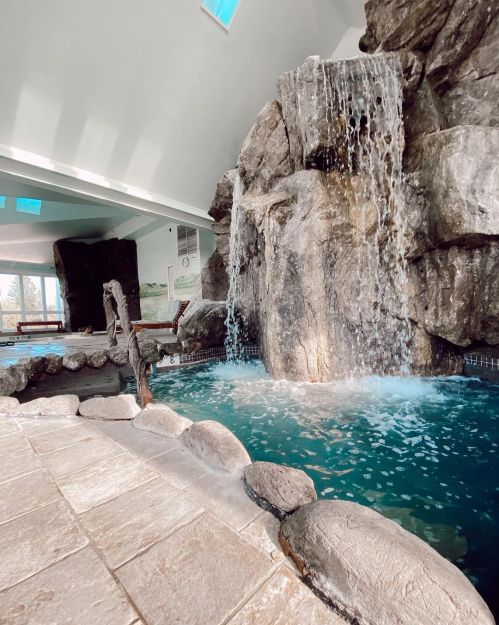 Indoor pool area featuring a rock waterfall, clear blue water, and stone pathways. Bright, airy atmosphere.