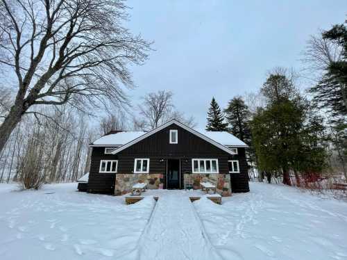 A dark wooden house with stone accents surrounded by snow and trees on a cloudy winter day.