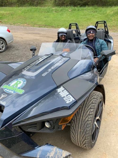Two people wearing helmets sit in a black three-wheeled vehicle on a dirt road, surrounded by greenery.