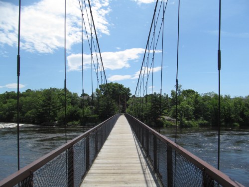 A suspension bridge spans a river, surrounded by lush greenery and a clear blue sky.