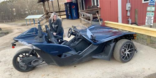 A woman stands next to a blue three-wheeled vehicle parked on a dirt path near a red building.