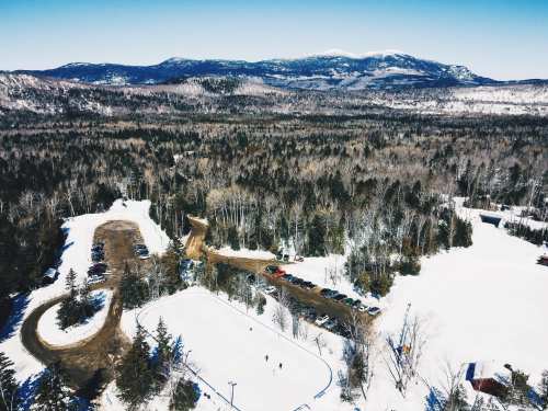 Aerial view of a snowy landscape with a parking area, winding road, and distant mountains under a clear blue sky.