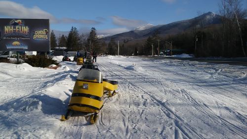 A snow-covered road with snowmobiles parked, surrounded by trees and mountains under a clear blue sky.