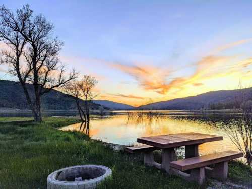 A serene lakeside scene at sunset, featuring a picnic table, fire pit, and trees against a colorful sky.