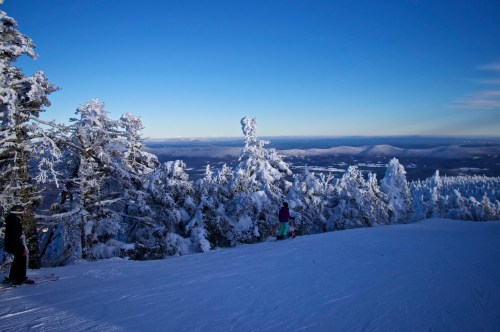 A snowy mountain landscape with trees covered in frost and a clear blue sky, featuring a skier in the foreground.