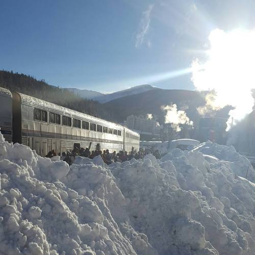 A train parked near a snowy landscape, with people gathered and mountains in the background under a bright sun.