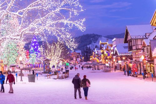 A snowy street lined with festive lights and decorations, with people walking hand in hand under a twilight sky.