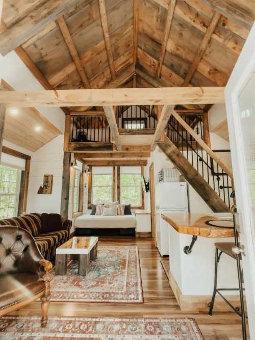 Cozy cabin interior with wooden beams, a sofa, a bed, and a staircase leading to a loft area. Warm, inviting atmosphere.