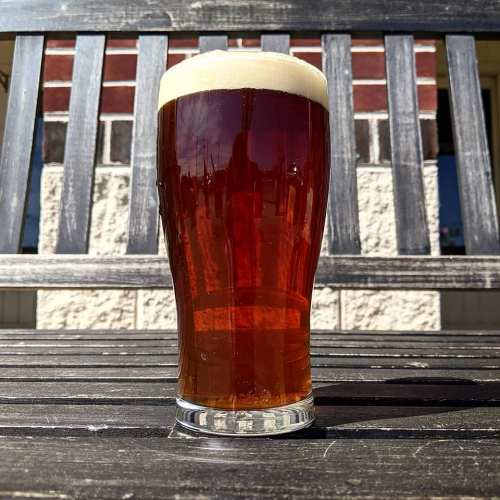 A glass of amber beer with a frothy head sits on a wooden table, with a blurred background of a building.