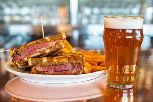 A plate with two grilled sandwiches filled with meat, served with fries, alongside a glass of amber beer.