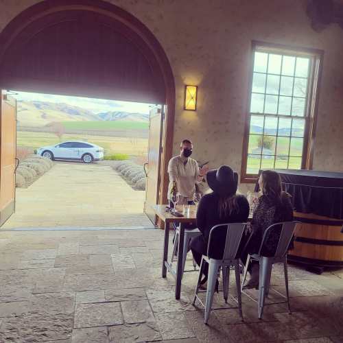 A server talks to two guests at a table inside a venue, with a scenic view of mountains and a car outside.