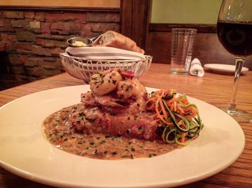 A plate of shrimp atop a savory sauce, accompanied by colorful vegetable noodles and a side of bread and wine.