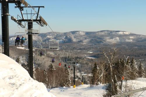 Ski lift chairs ascend a snowy mountain, surrounded by trees and distant hills under a clear blue sky.