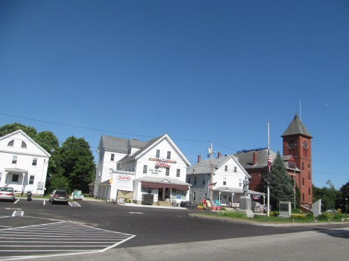 A small town scene featuring white buildings, a clock tower, and clear blue skies. Cars are parked in the foreground.