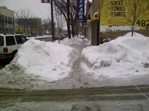 Snow piles line a sidewalk in a city, with a few trees and buildings visible in the background on a snowy day.