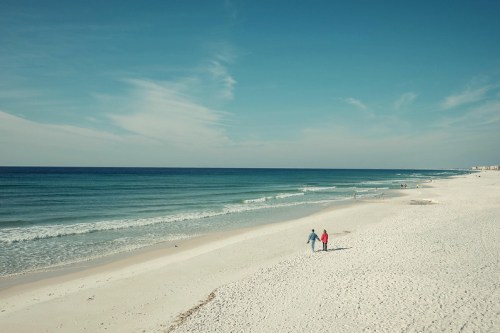A couple walks hand in hand along a serene beach with gentle waves and a clear blue sky.