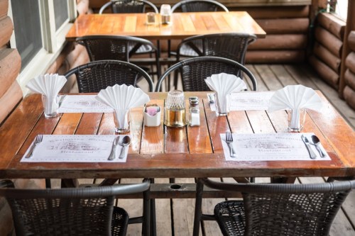A rustic wooden dining table set with folded napkins, menus, and condiments, surrounded by wicker chairs.
