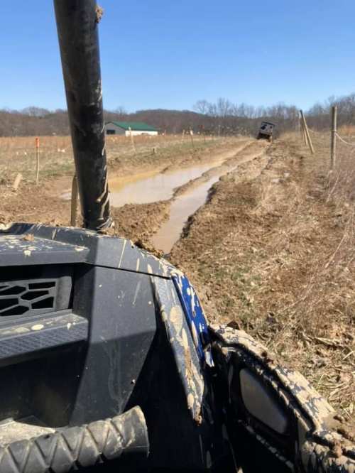 A muddy off-road trail with tire tracks and puddles, viewed from the front of an ATV. Trees and hills in the background.