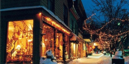 A snowy street at night, lined with shops adorned with warm lights and festive decorations.