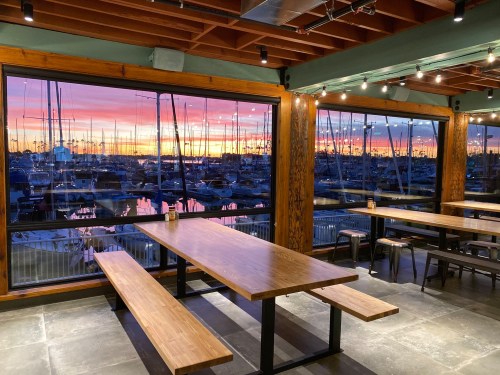 A cozy restaurant interior with wooden tables, overlooking a marina at sunset with vibrant colors in the sky.