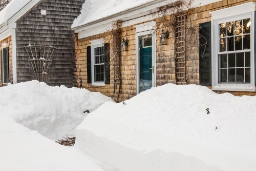 A snow-covered house with wooden siding, featuring a blue door and windows, surrounded by large snowdrifts.