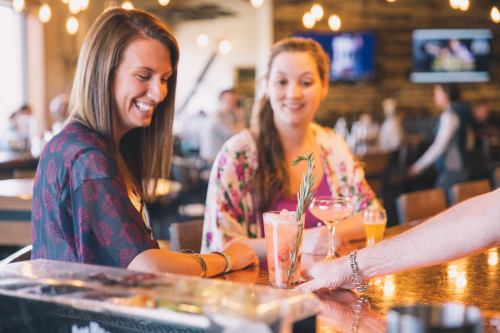 Two women smile at a bar while a bartender serves a drink with a sprig of rosemary. Warm, inviting atmosphere.