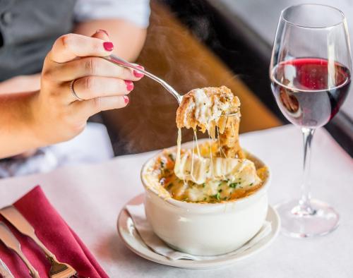 A hand holding a spoon with cheesy soup, next to a glass of red wine on a table.
