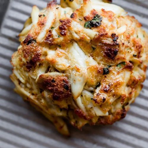 A golden-brown crab cake with a crispy exterior, featuring bits of crab and herbs, resting on a textured surface.