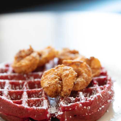 A red velvet waffle topped with crispy fried chicken and drizzled with syrup, served on a white plate.