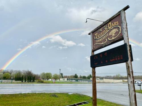 A wooden sign for Twisted Oaks, Tennessee, with a vibrant rainbow in the background after a rainstorm.