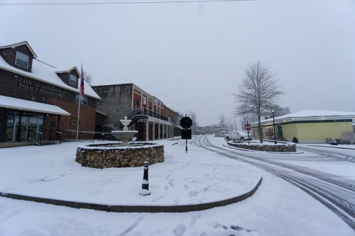 Snow-covered street scene with buildings, a fountain, and a stop sign, creating a quiet winter atmosphere.