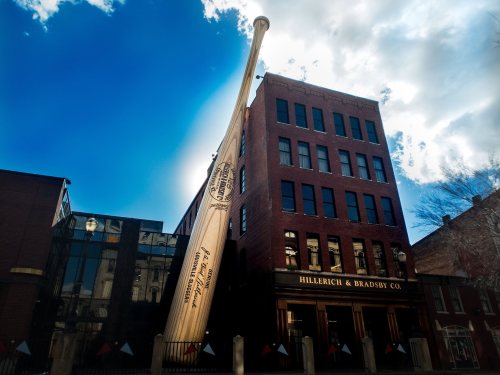 A large baseball bat leans against the Hillerich & Bradsby Co. building under a bright blue sky.