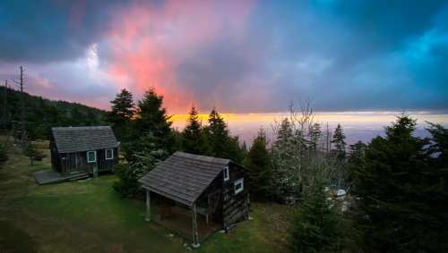Two rustic cabins surrounded by trees, with a colorful sunset and dramatic clouds in the background.